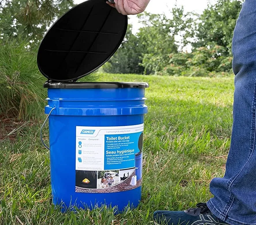 Blue colored bucket portable camping toilet for trucks, placed in a camping ground, lid being opened by a man.